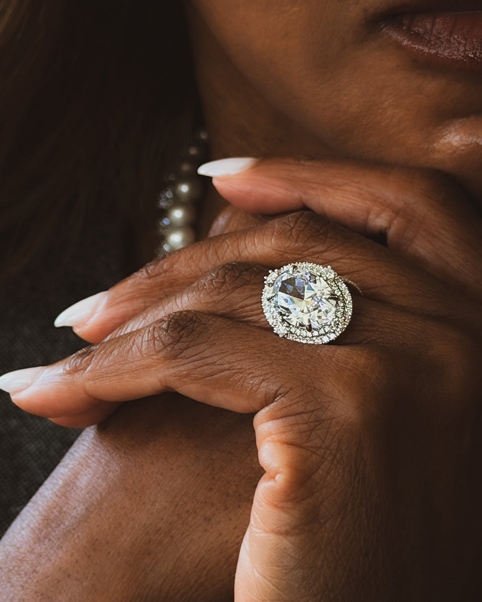 A large round diamond ring with a double halo of pavé diamonds in white gold, worn on the hand of a woman resting her chin on her knuckles. Pearl jewelry is visible in the soft-focus background.